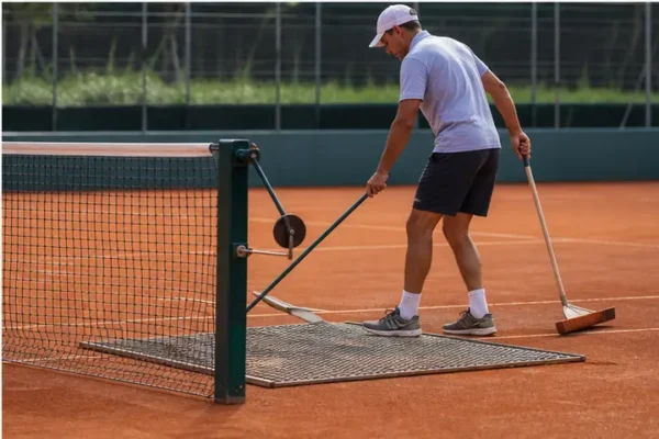 Manutentore utilizza stuoia e spazzola su un campo da tennis in terra battuta.